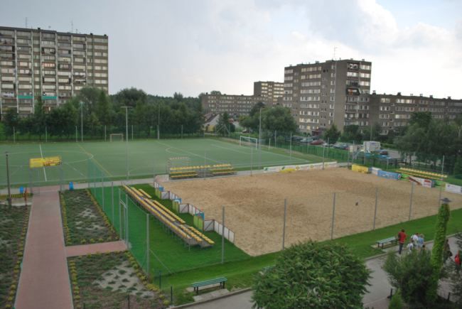 widok na duże boisko trwaiaste do piłki nożnej, boisko do siatkowki plazowej pokryte piaskiem z trybunami do siedzenia  dookoła bloki oś Niepodległosci , po lewej stronie zagodposarowany zielony teren