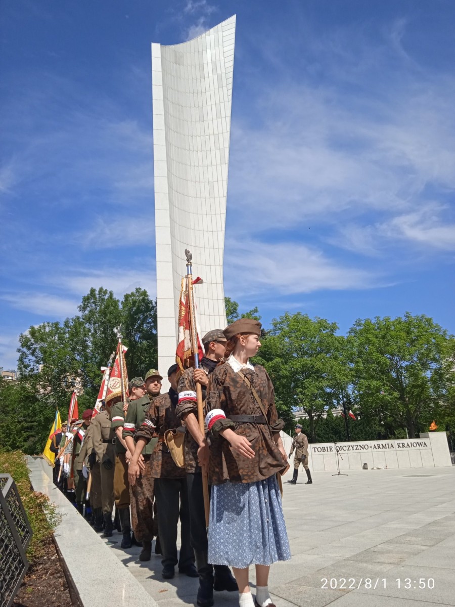 Przebrani za powstańców warszawskich młodzi ludzie stoją na baczność przy pomniku 