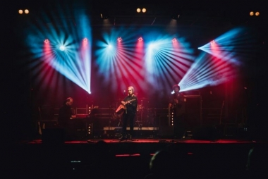 Trzech muzyków na scenie oświetlonej w kolorze czerwono-niebieskim . Dwóch grających na gitarach jeden na klawiszach. 
