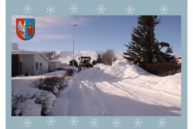 pług odśnieżajacy bardzo zaśnieżony teren wokół hałdy sniegu , niebo niebieskie po lewej stronie małe białe domy  po prawej stronie drzewa iglaste wyłaniają się ze śniegu 