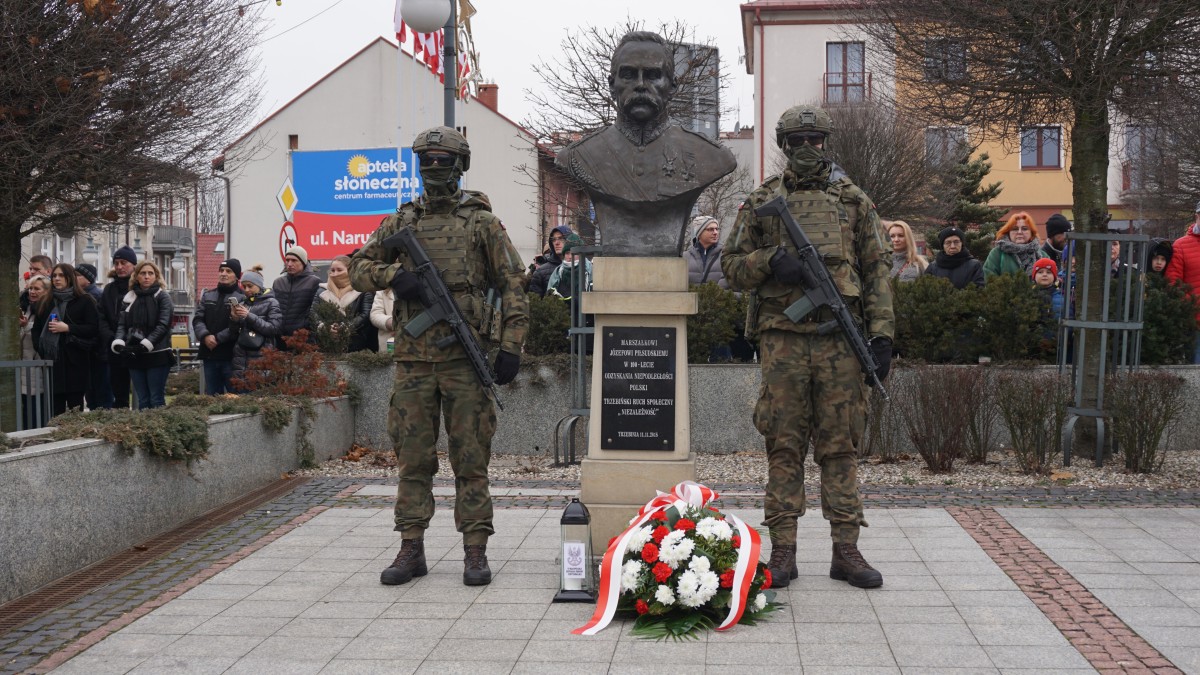 Zdjęcie. Uroczysta warta dwóch żołnierzy przy popiersiu Marszałka Józefa Piłsudskiego. Przed popiersiem leżą złożone kwiaty. W tle widać obsewatorów wydarzenia,