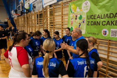 Zdjęcie zespołu dziewcząt w piłkę ręczną na hali sportowej. Dziesięć zawodniczek stoi w kręgu obok swojego trenera. 