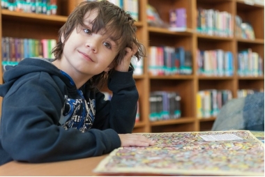 Zdjęcie młodego chłopca w bibliotece. W tyle regały z książkami. Przed chłopcem otwarta książka. 